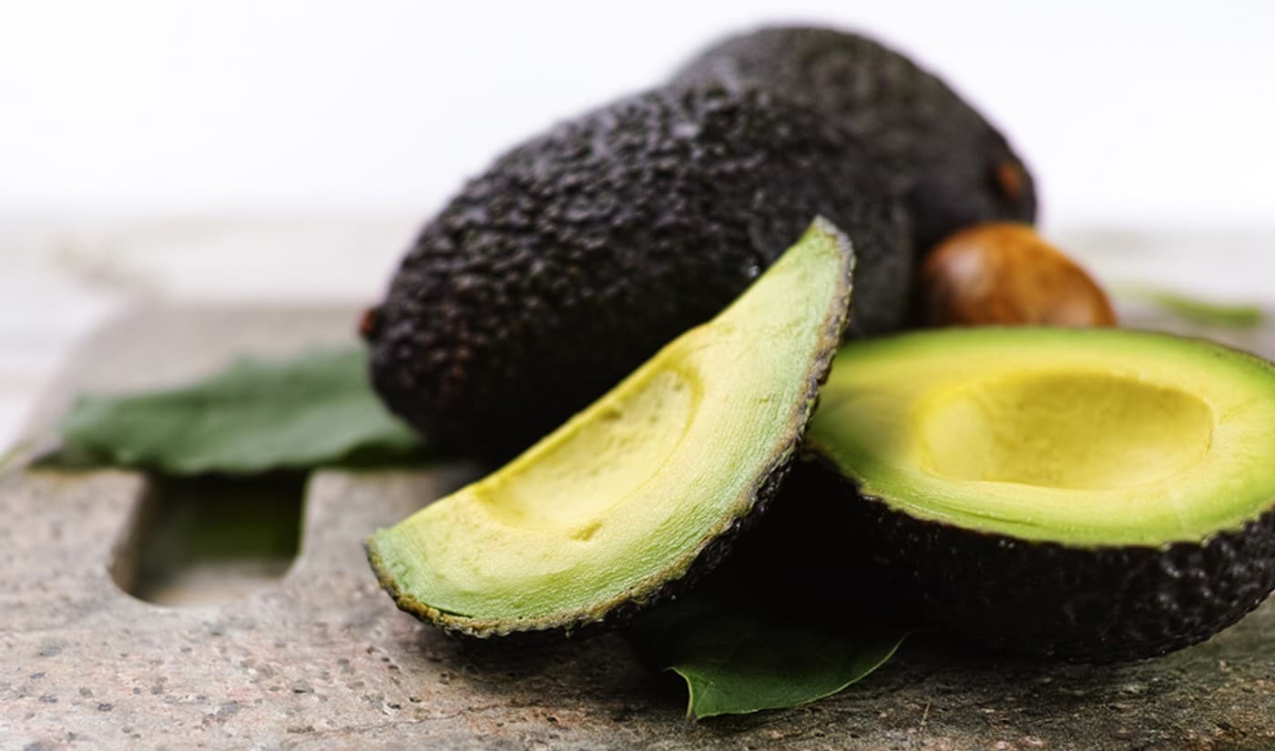 Aguacates de piel oscura, uno de ellos abierto por la mitad, sobre una superficie de piedra con hojas verdes