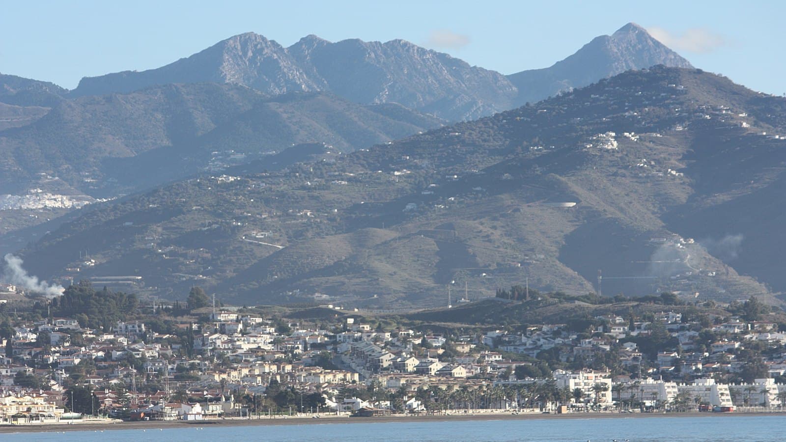 Vista desde el mar de la costa de Algarrobo: una franja de casas y bloques blancos junto a la playa, lomas con cultivos y viviendas dispersas detrás y, al fondo, las cumbres rocosas de la sierra bajo un cielo despejado.
