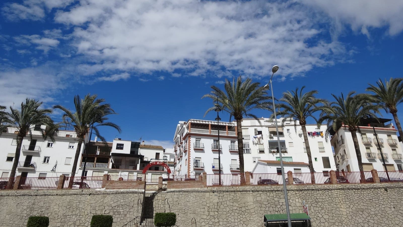Edificios blancos de varias plantas con balcones en Algarrobo, tras una hilera de palmeras y una barandilla roja sobre un muro de piedra, bajo un cielo azul con nubes.