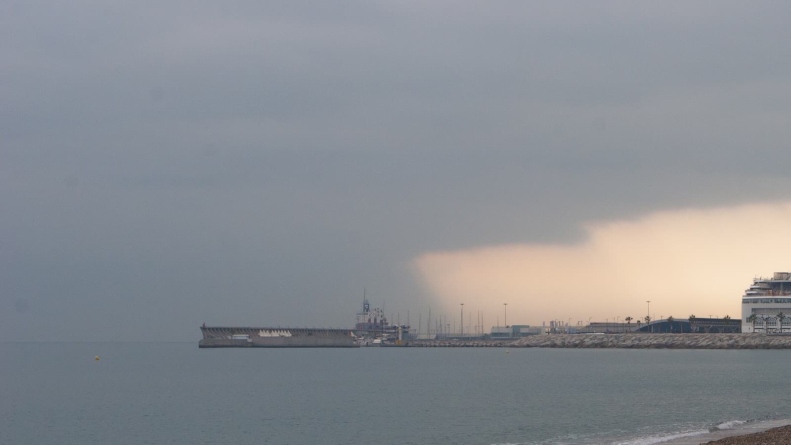 Un frente de nubes oscuras y lluvia avanza sobre el mar hacia el puerto de Málaga, con el dique y un crucero al fondo.