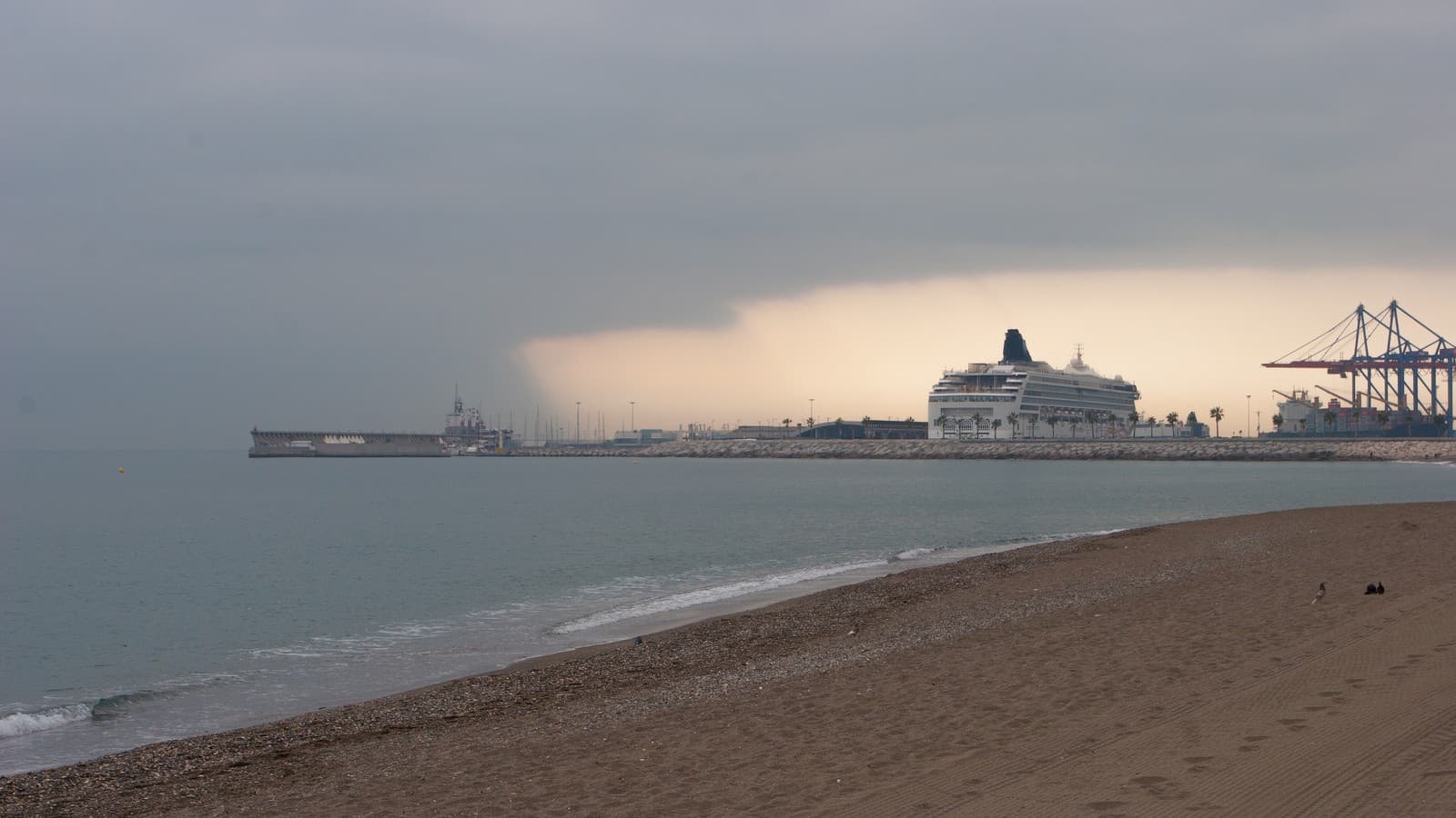 Un frente de nubes oscuras y lluvia avanza sobre el mar hacia el puerto de Málaga, con el dique y un crucero al fondo.
