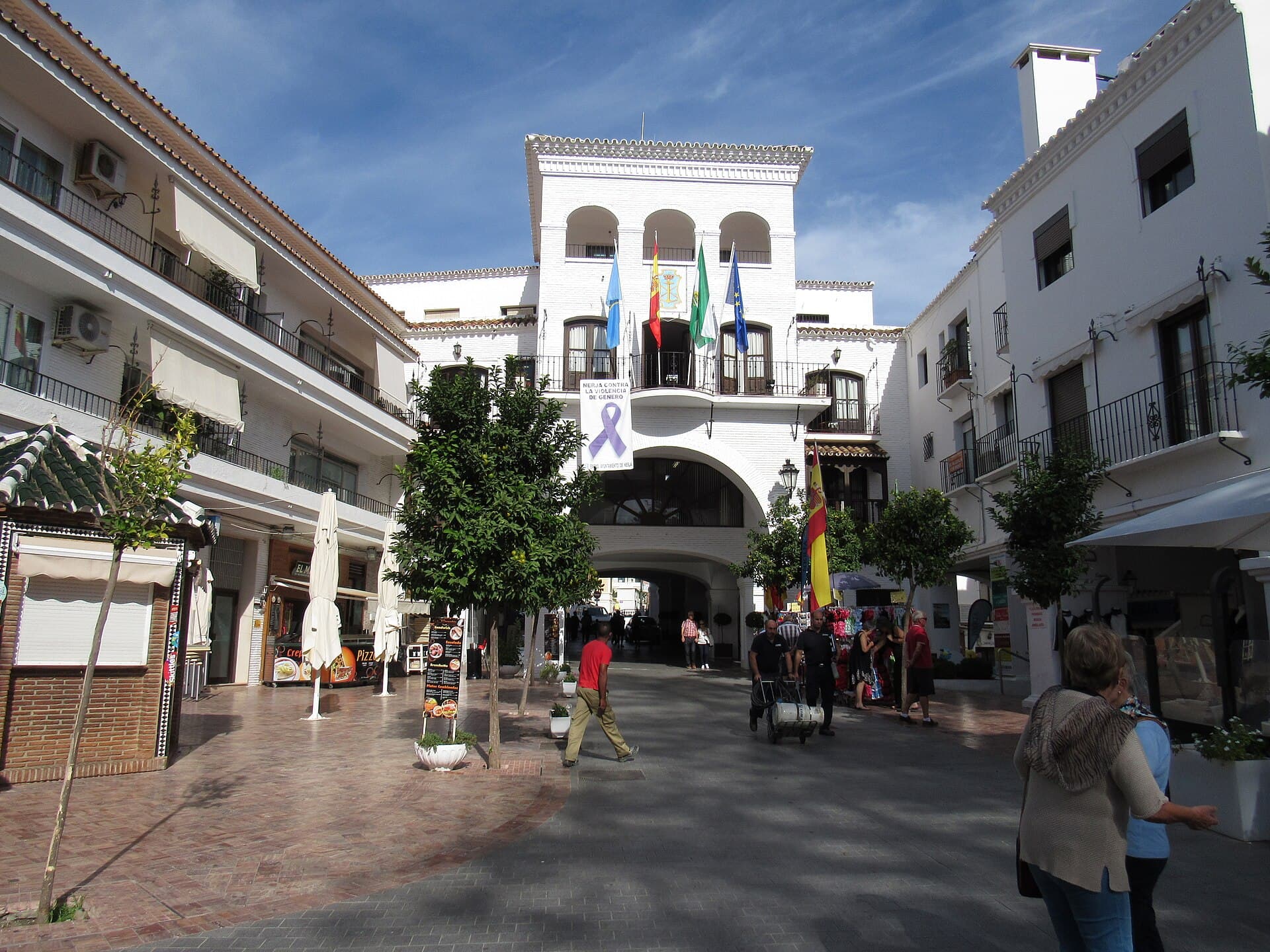 Fachada blanca del Ayuntamiento de Nerja, con arcos, balcón y banderas, vista desde la plaza peatonal con naranjos y vecinos paseando.