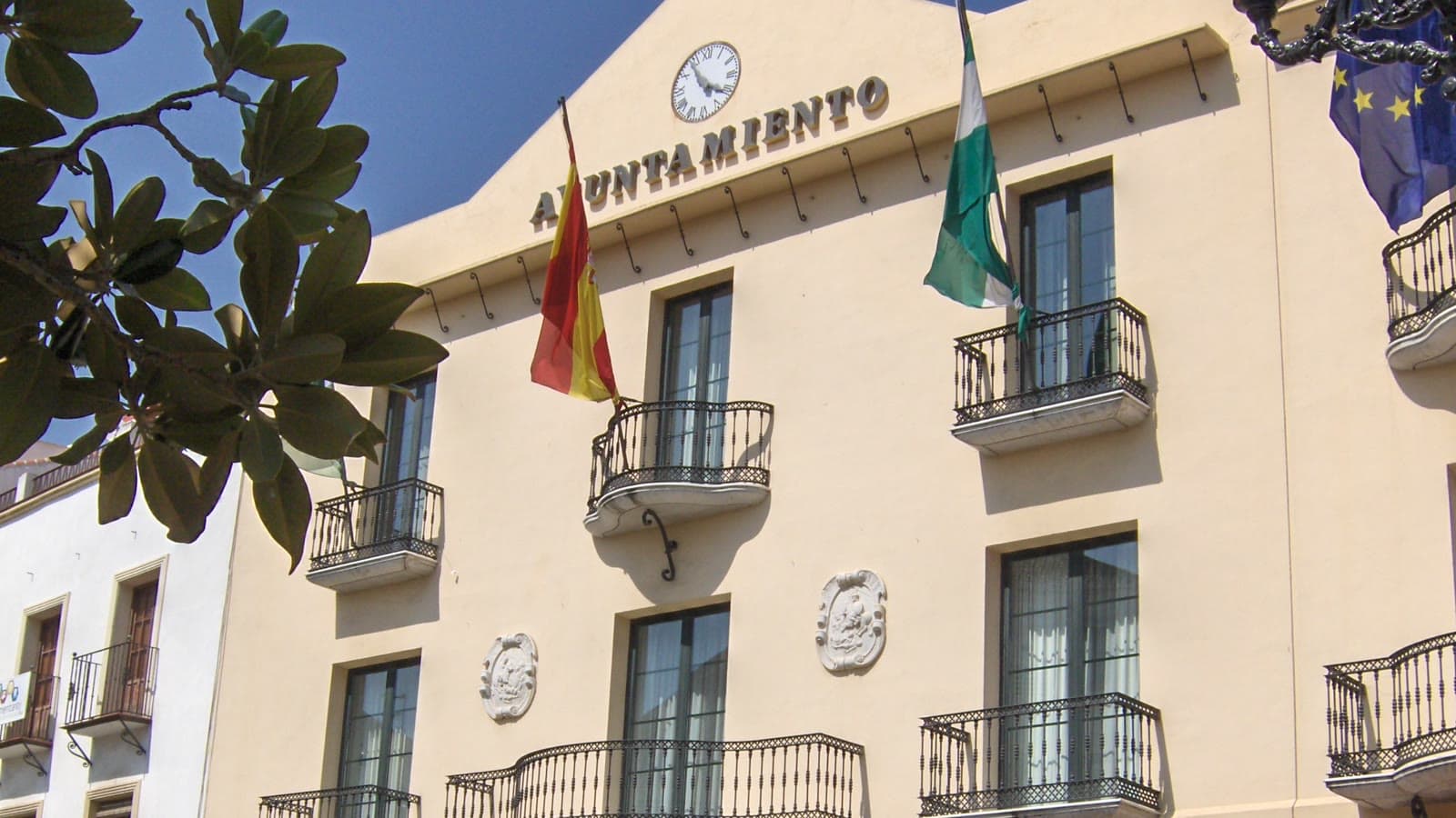 Fachada del Ayuntamiento de Vélez-Málaga, de color crema, con el rótulo «Ayuntamiento», un reloj, balcones de forja y las banderas de España, Andalucía y la Unión Europea.