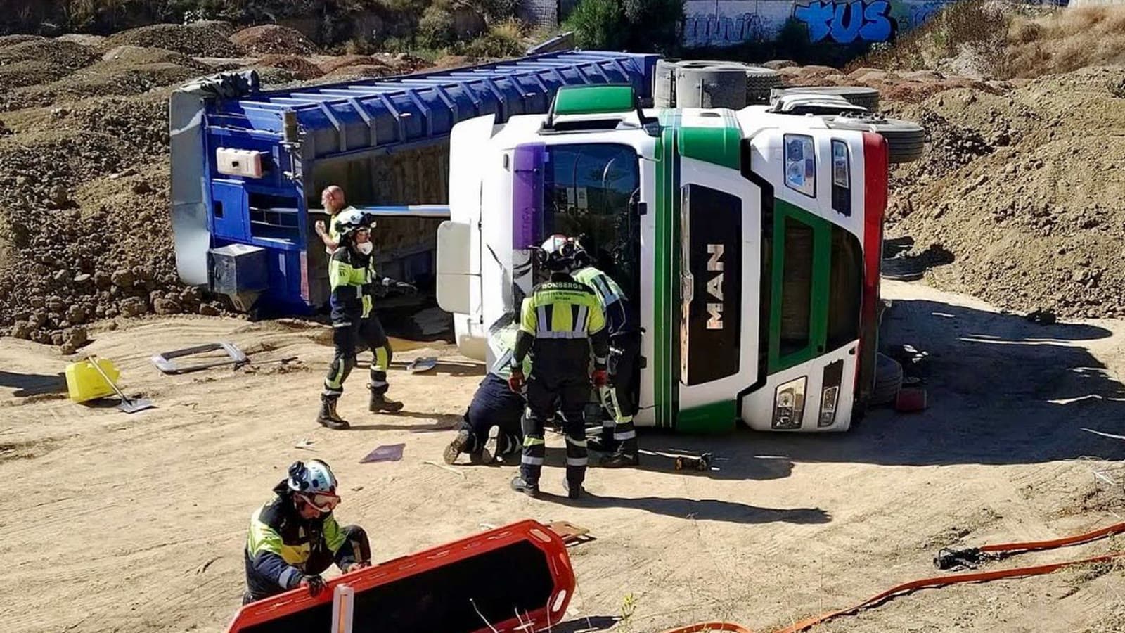 Bomberos del Consorcio Provincial de Málaga trabajan junto a la cabina de un camión blanco y verde volcado de lado sobre un terreno de tierra; en primer plano, un bombero prepara una camilla de rescate naranja.