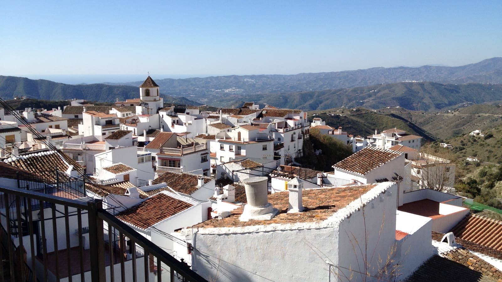 Vista de Canillas de Aceituno desde una baranda: casas blancas con tejados de teja, el campanario de la iglesia y, al fondo, los montes de la Axarquía y el mar.
