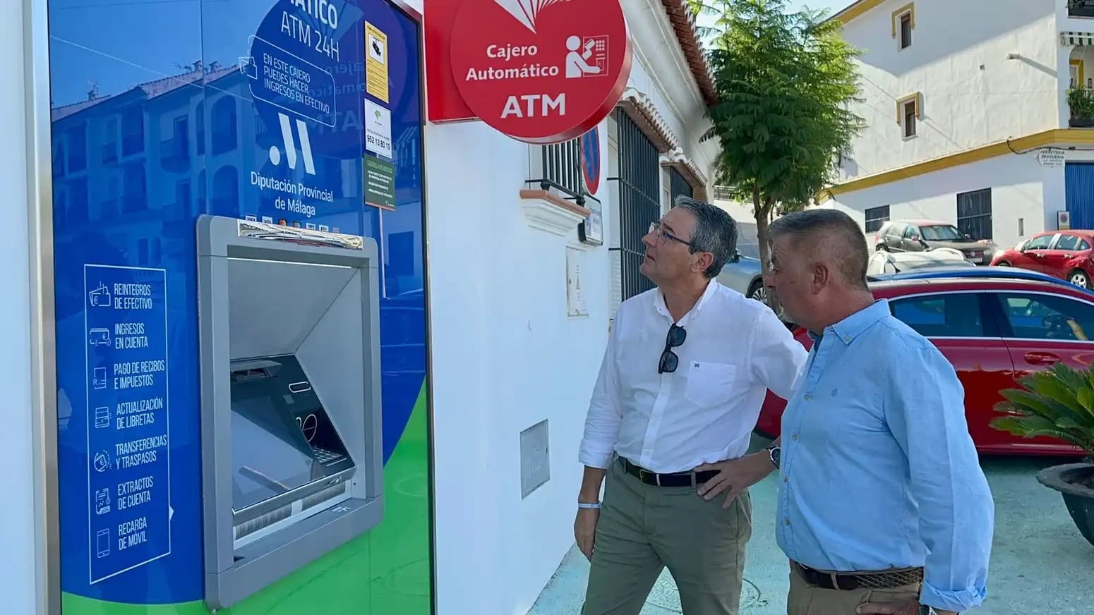 Dos hombres miran un cajero automático azul y verde con el logotipo de la Diputación Provincial de Málaga, empotrado en la fachada blanca de un edificio de Canillas de Albaida, bajo un rótulo rojo de «Cajero automático ATM»