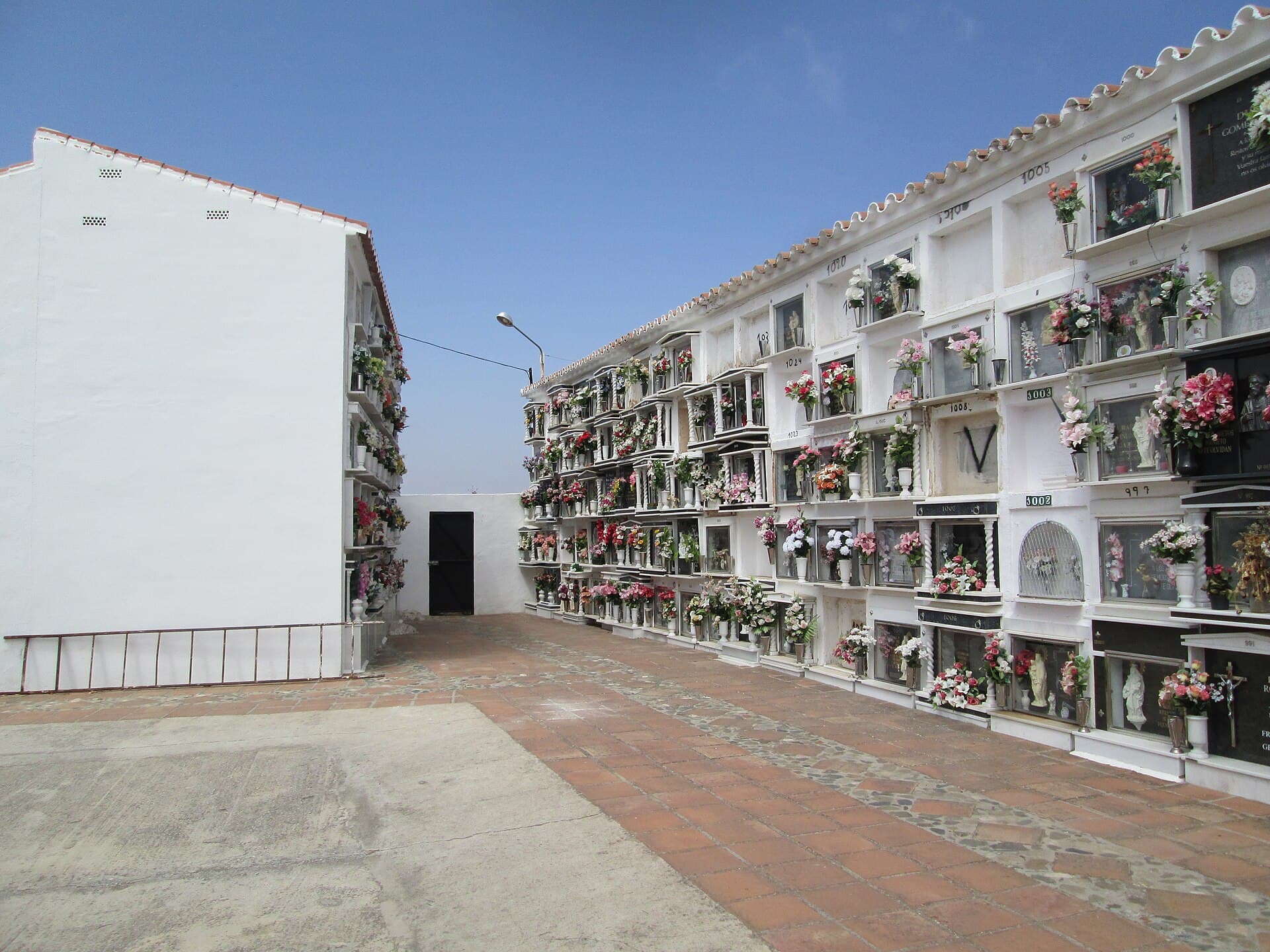 Pasillo del cementerio de Comares, con paredes blancas de nichos adornados con flores bajo un cielo azul.