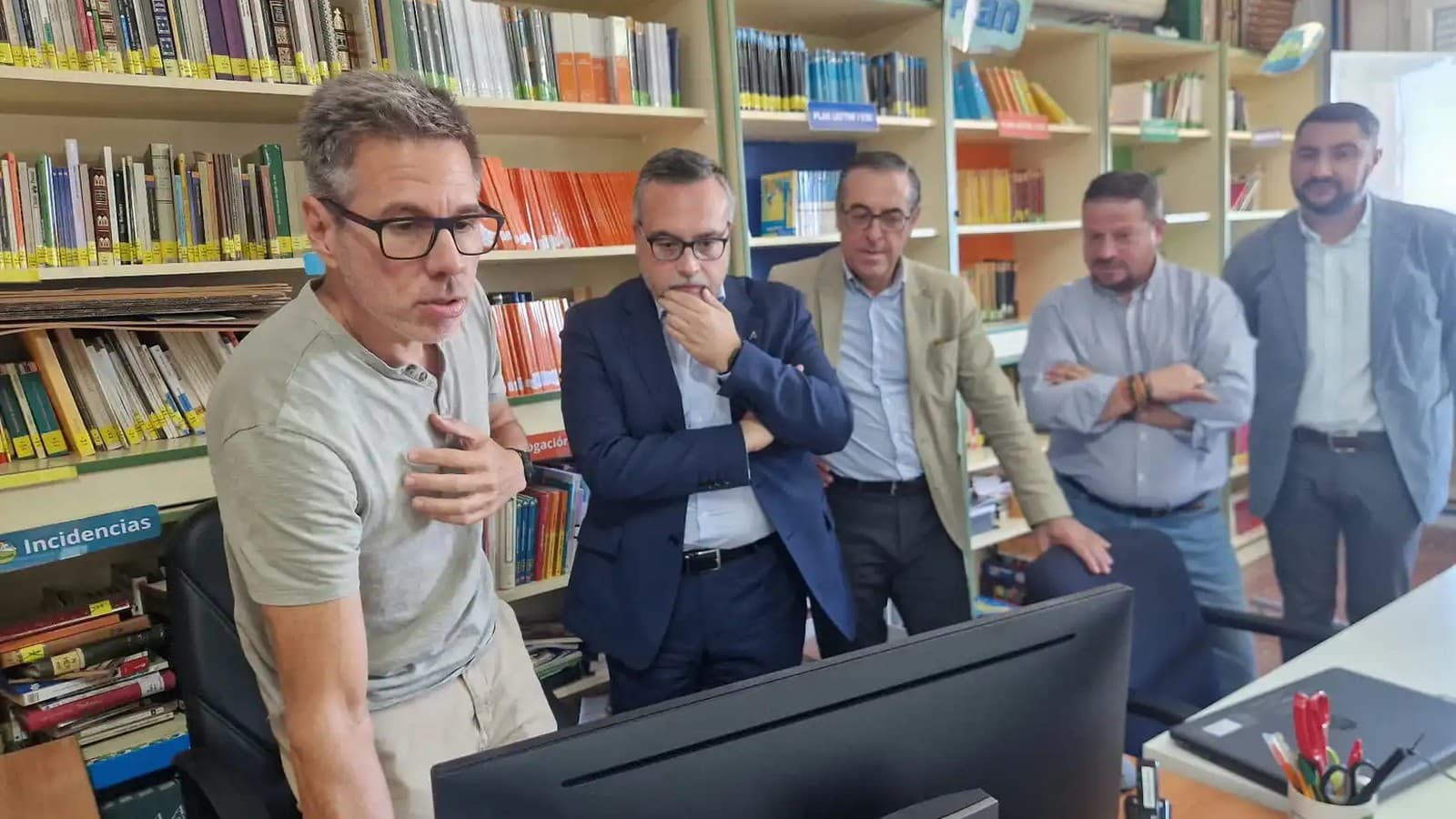 Un hombre con gafas y camiseta gris explica algo ante un ordenador a cuatro hombres que le escuchan de pie, en una biblioteca escolar con estanterías llenas de libros.