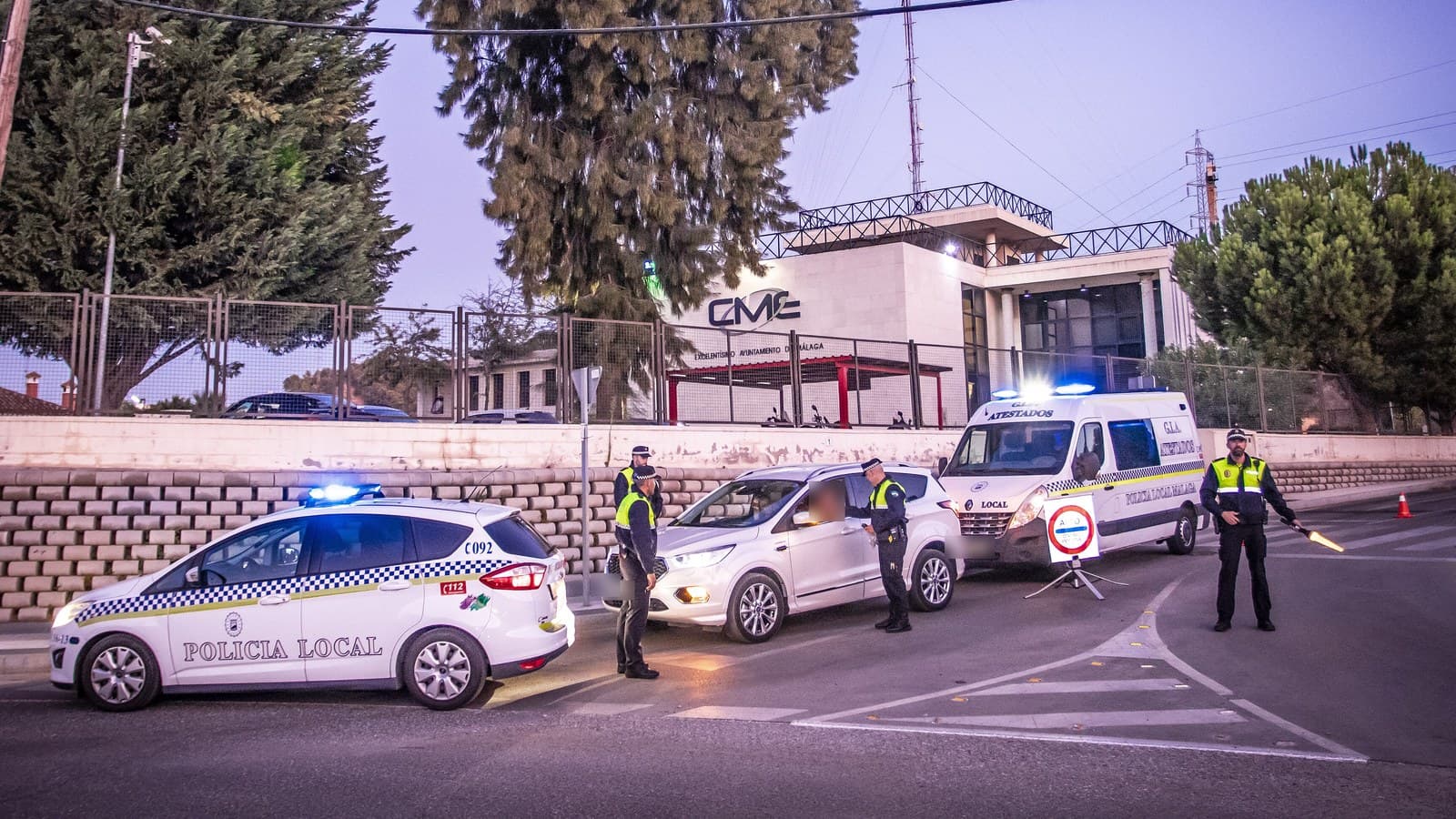 Control de tráfico de la Policía Local al anochecer: un coche patrulla con las luces encendidas, agentes con chaleco reflectante junto a un turismo detenido y un furgón de atestados al fondo.