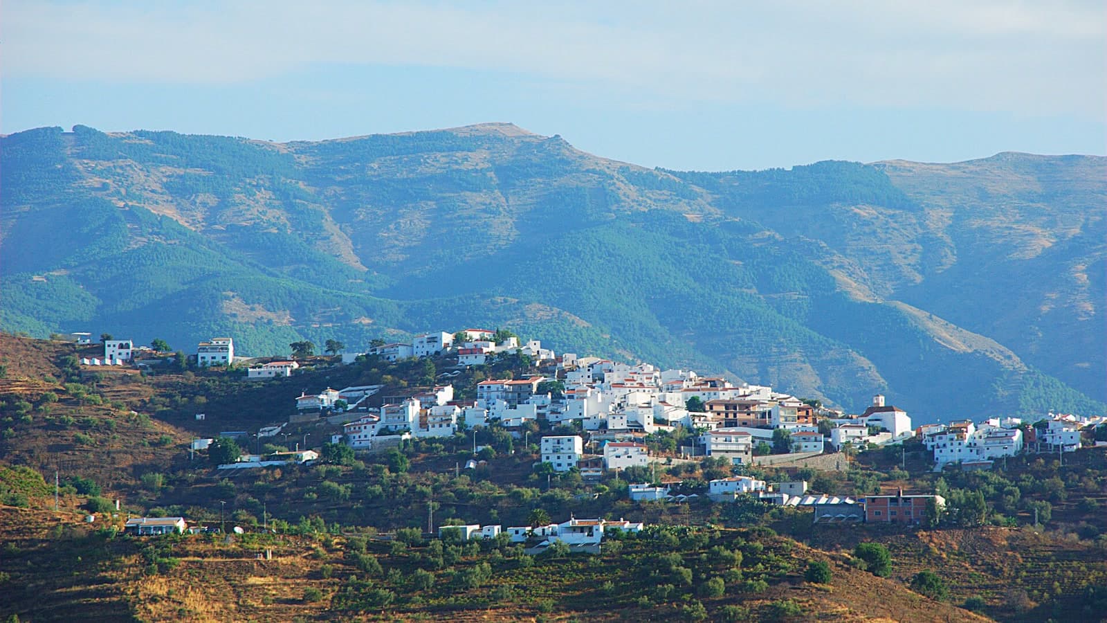 Caserío blanco de Corumbela sobre una ladera con árboles, con montañas al fondo, visto desde Sayalonga.