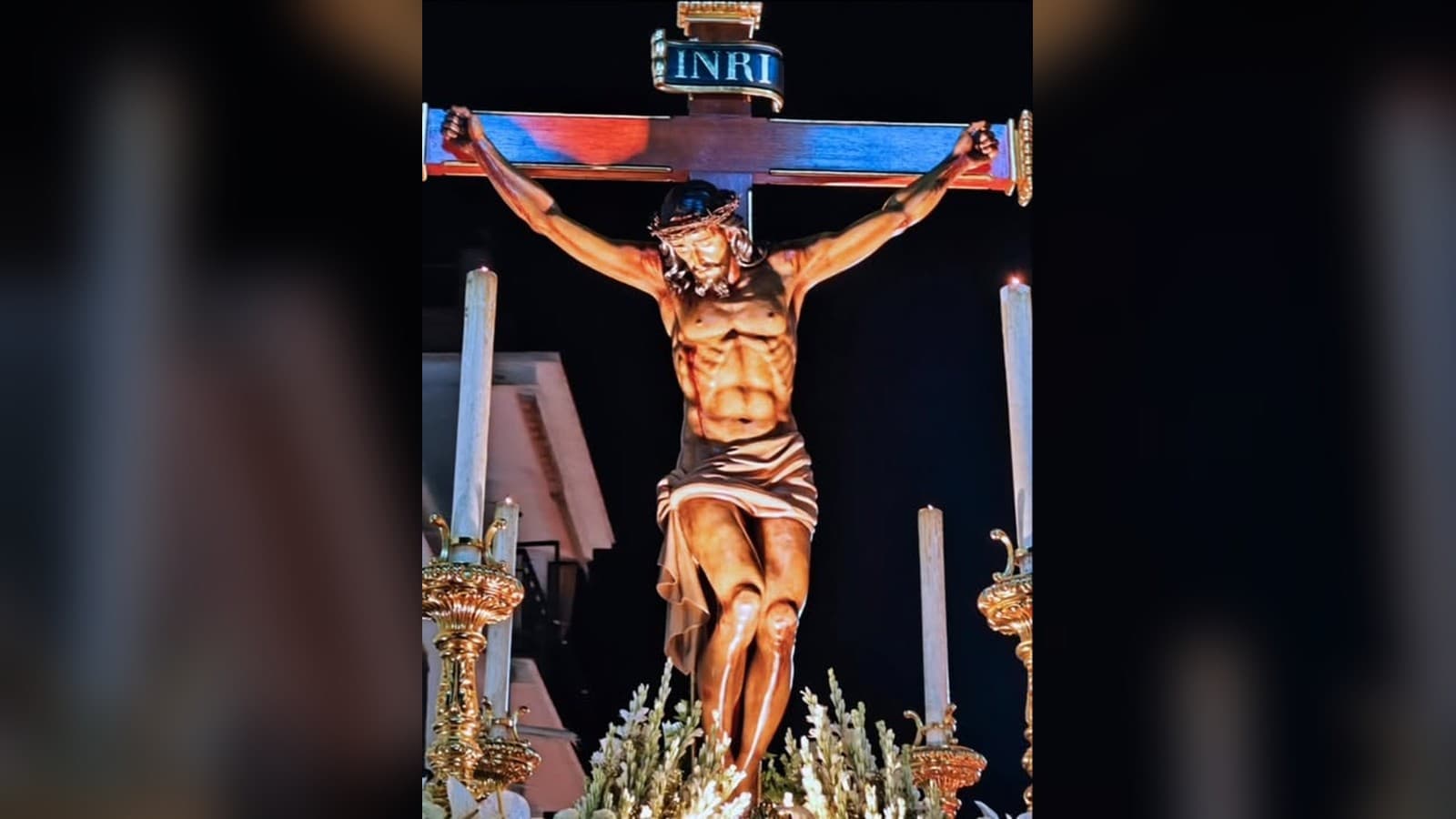 La imagen del Santísimo Cristo de la Salud de Benamocarra, crucificado, de noche, sobre su trono con flores blancas, cirios y candelabros dorados.