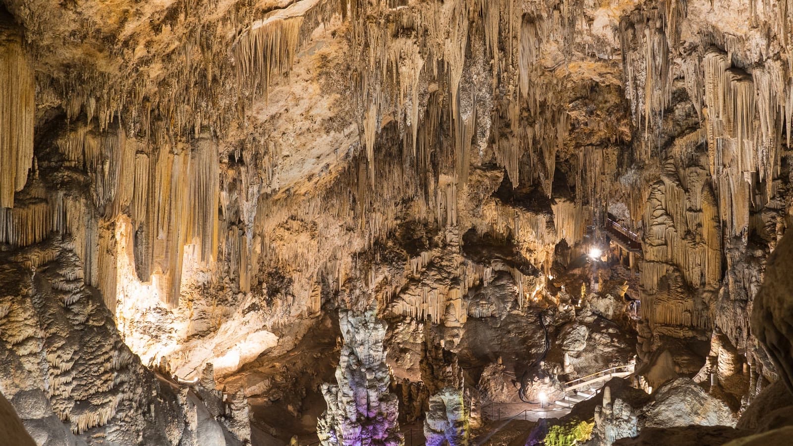 Interior de la Cueva de Nerja con estalactitas y columnas iluminadas en tonos ocres y una pasarela con barandilla al fondo.