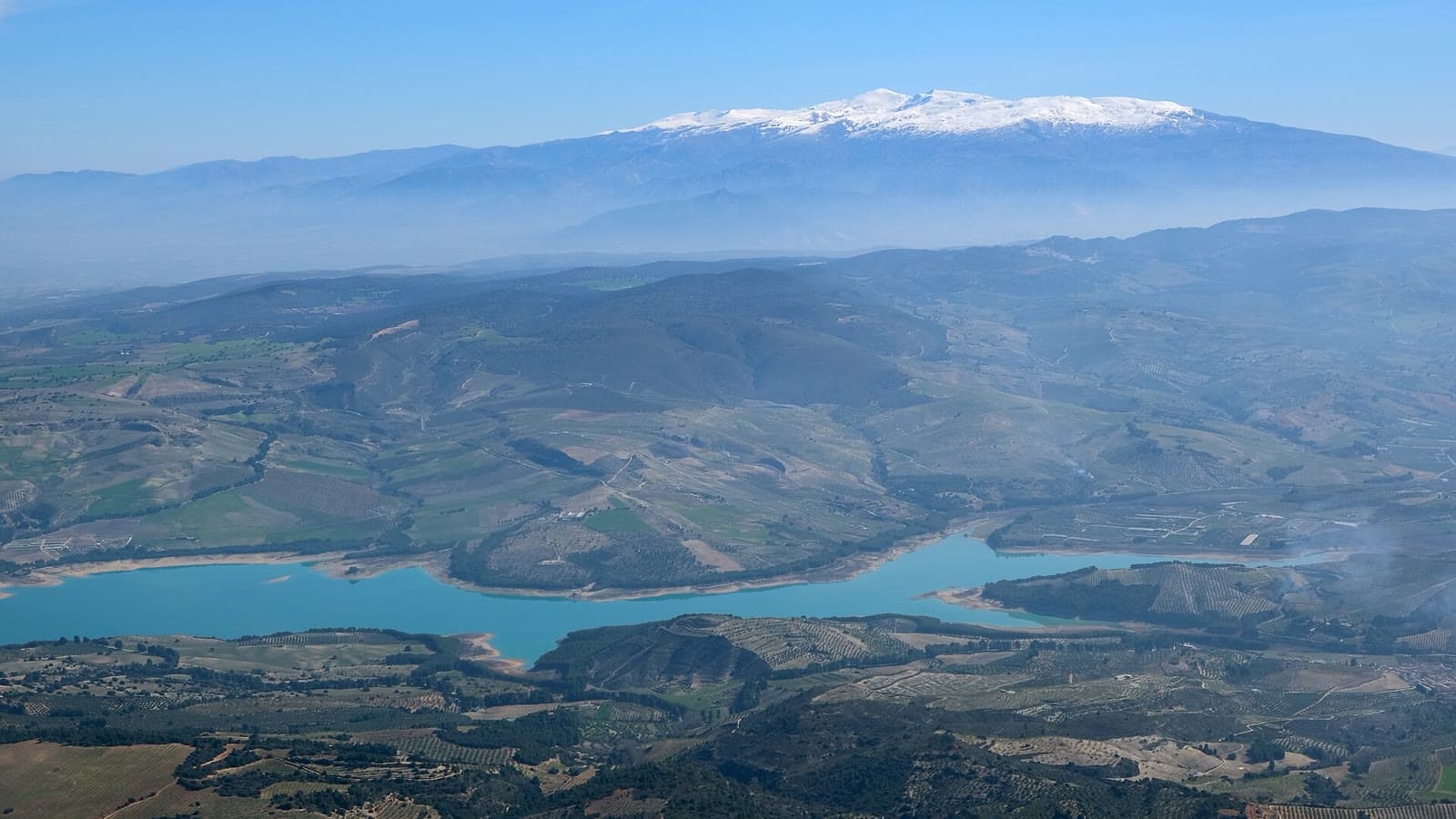 Vista aérea del embalse de La Viñuela, de color turquesa, entre lomas de olivar, con Sierra Nevada nevada al fondo.