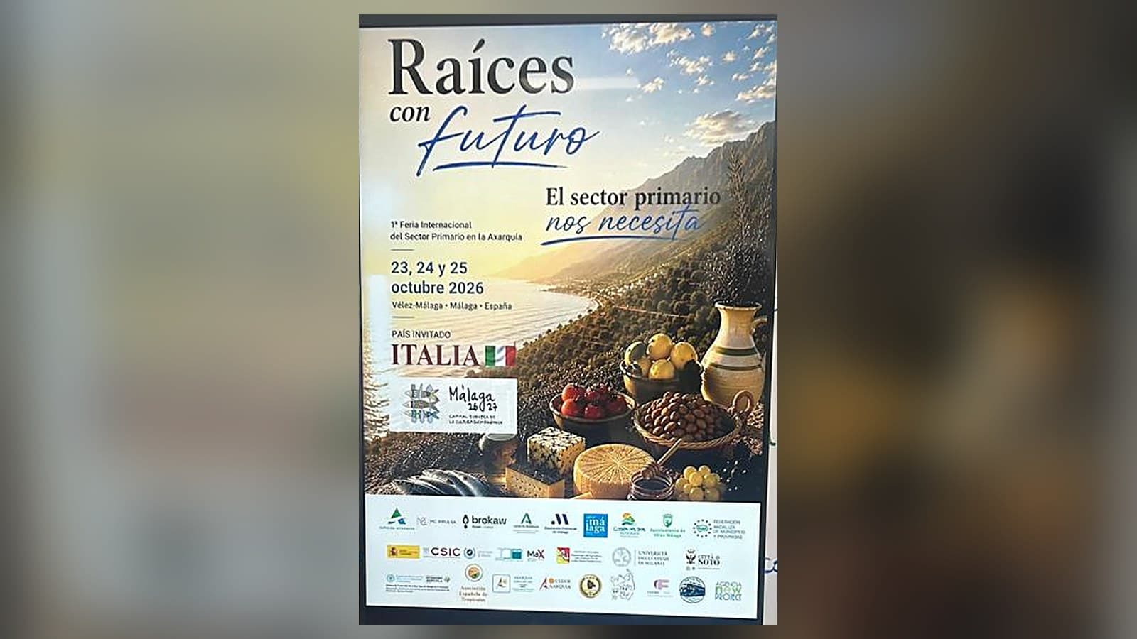 Cartel de FIAX «Raíces con futuro. El sector primario nos necesita»: 1.ª Feria Internacional del Sector Primario en la Axarquía, 23, 24 y 25 de octubre de 2026 en Vélez-Málaga, con Italia como país invitado, sobre una vista de la costa con un bodegón de quesos, frutas, aceitunas y una jarra.
