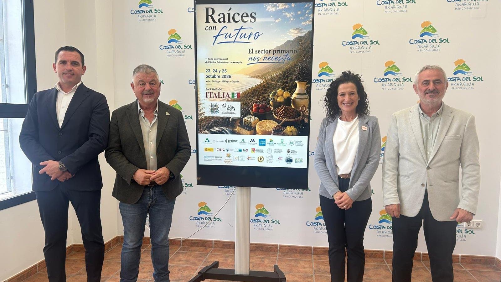 Cuatro personas posan junto a una pantalla con el cartel de la feria «Raíces con futuro», que anuncia la 1.ª Feria Internacional del Sector Primario de la Axarquía del 23 al 25 de octubre de 2026 con Italia como país invitado, ante un panel con el logotipo de la Mancomunidad Costa del Sol Axarquía.