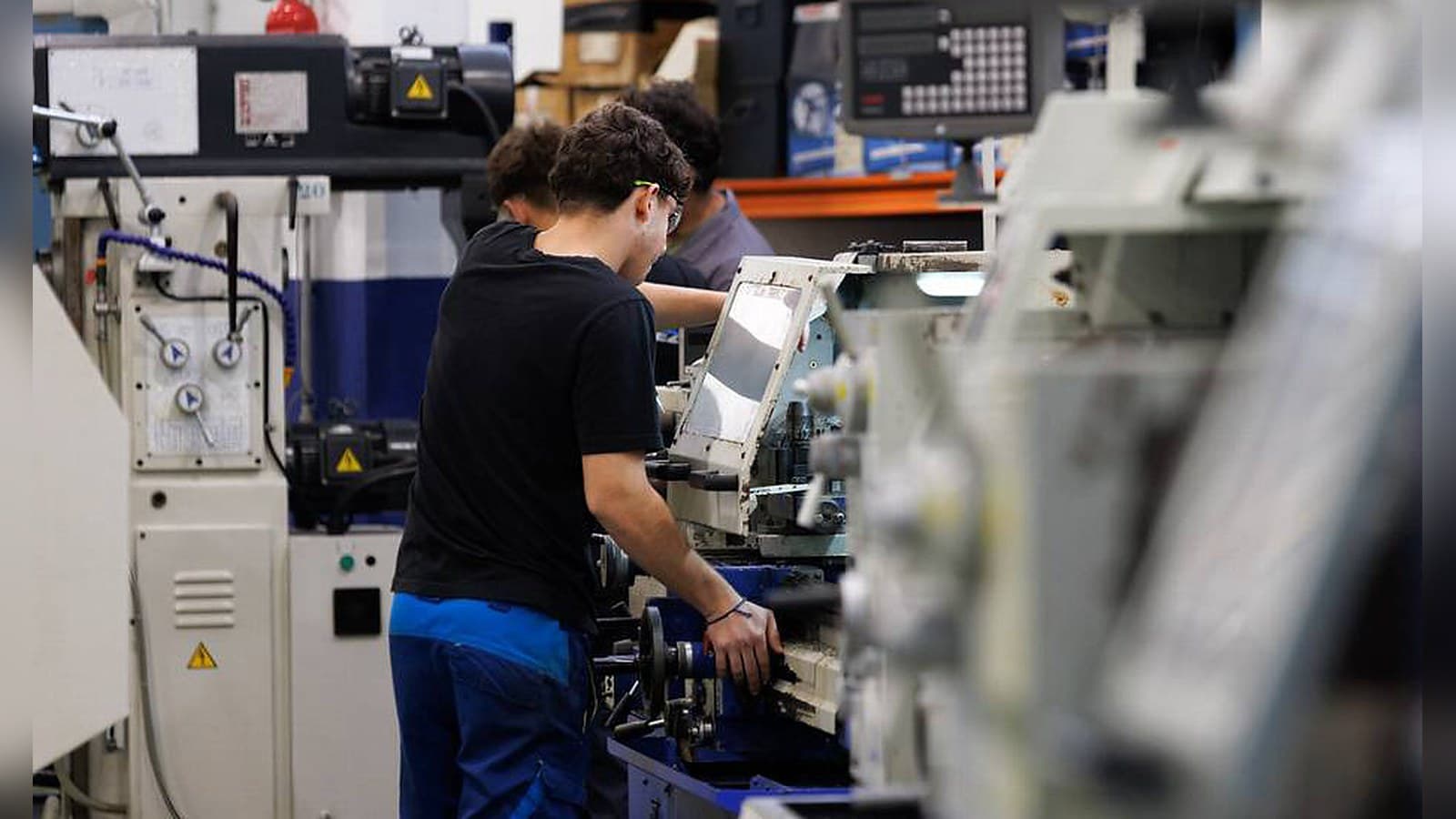 Un estudiante de Formación Profesional con camiseta negra, pantalón azul de trabajo y gafas de protección maneja un torno en un taller, con otros alumnos detrás y máquinas desenfocadas en primer plano
