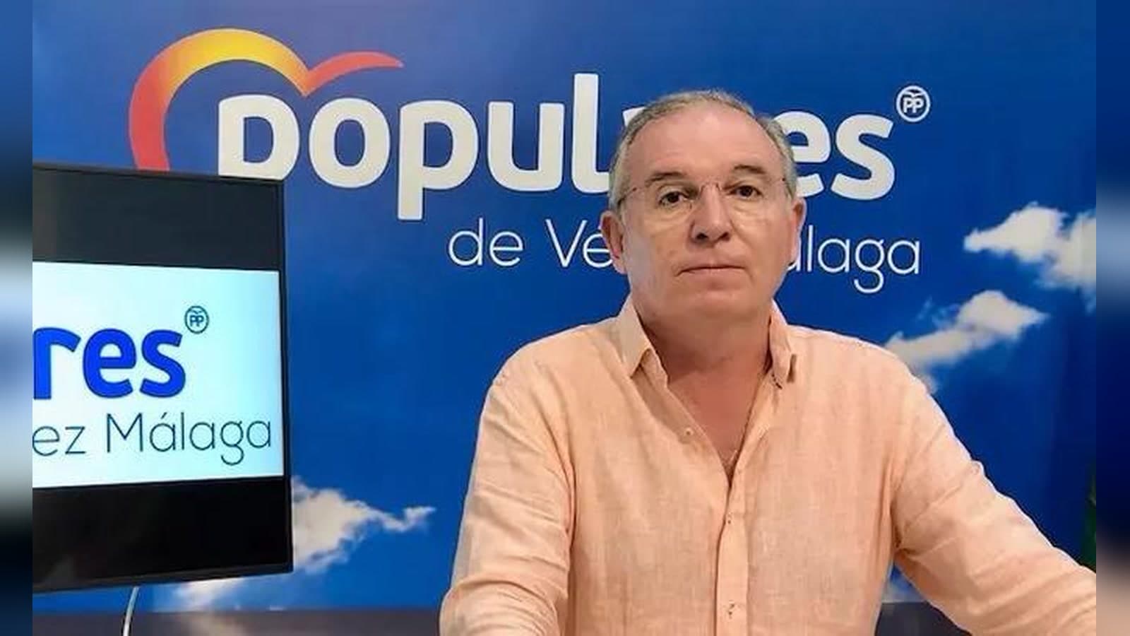 Francisco Delgado Bonilla, con camisa clara y gafas, ante un fondo azul con el rótulo «Populares de Vélez-Málaga».