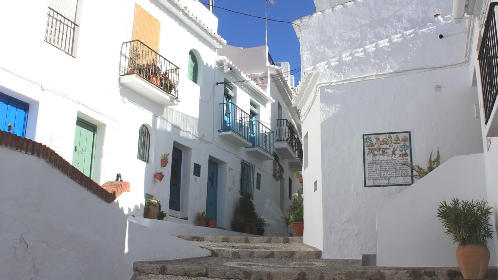 Calle empedrada con escalones en el casco antiguo de Frigiliana, entre casas encaladas con balcones de hierro, macetas y un panel de azulejos en la pared.