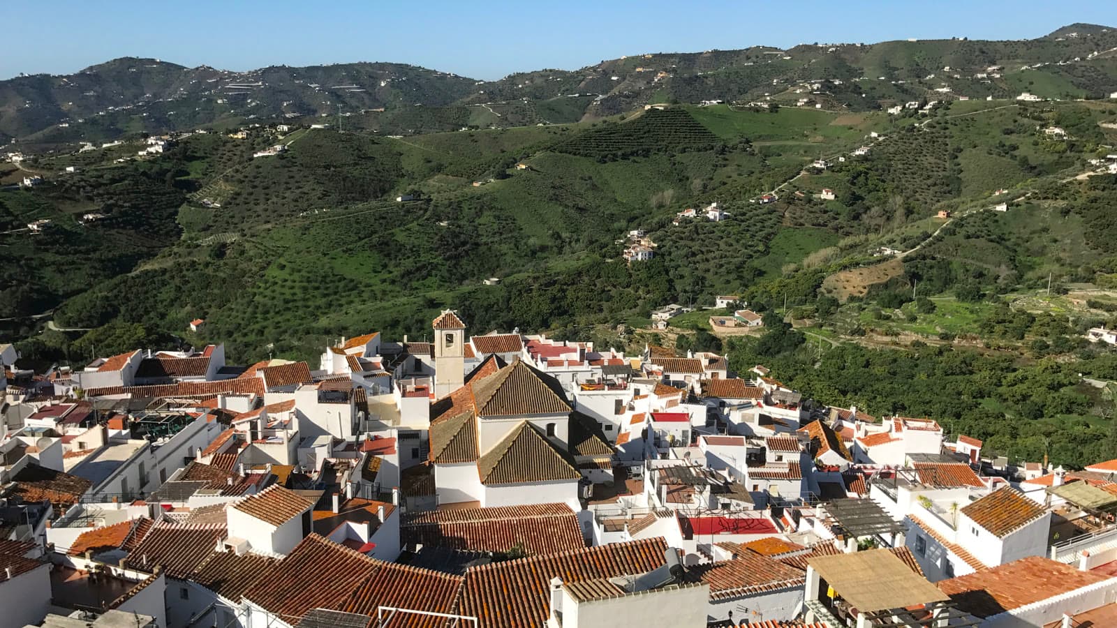Vista desde lo alto del casco urbano de Frigiliana, con tejados de teja y la torre de la iglesia, y al fondo lomas verdes salpicadas de casas blancas.