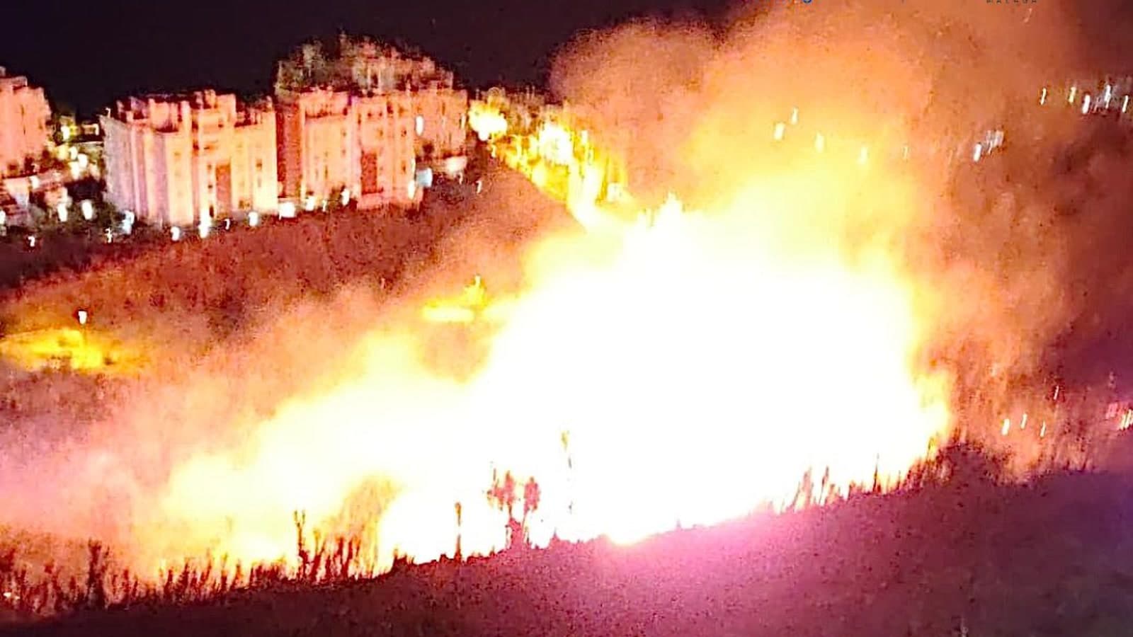 De noche, grandes llamas y humo en una ladera con matorral, con los edificios iluminados de una urbanización al fondo.