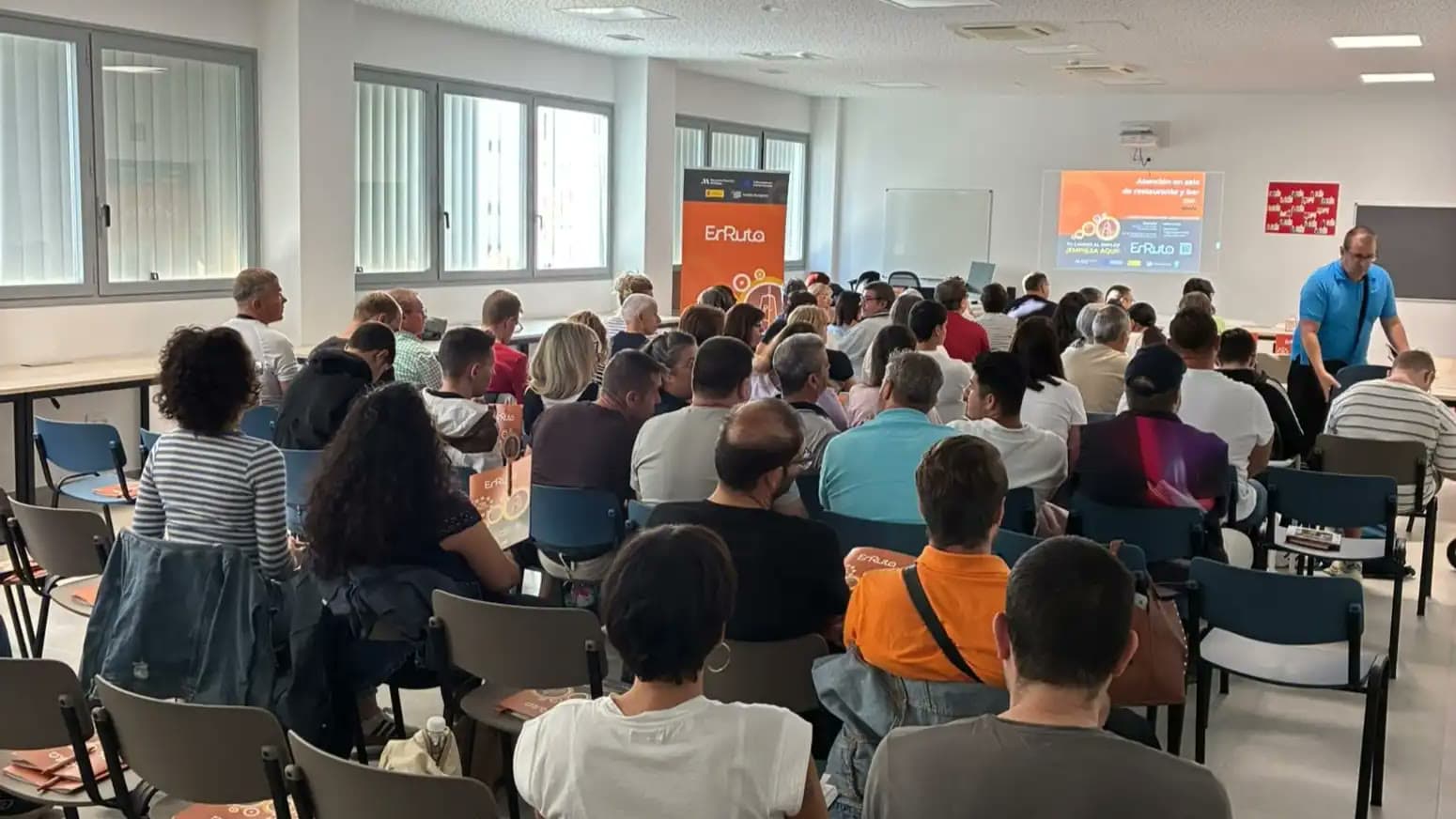 Decenas de personas, vistas de espaldas, sentadas en un aula del Centro de Formación y Empleo de Nerja frente a una pantalla y un expositor naranja del programa EnRuta.