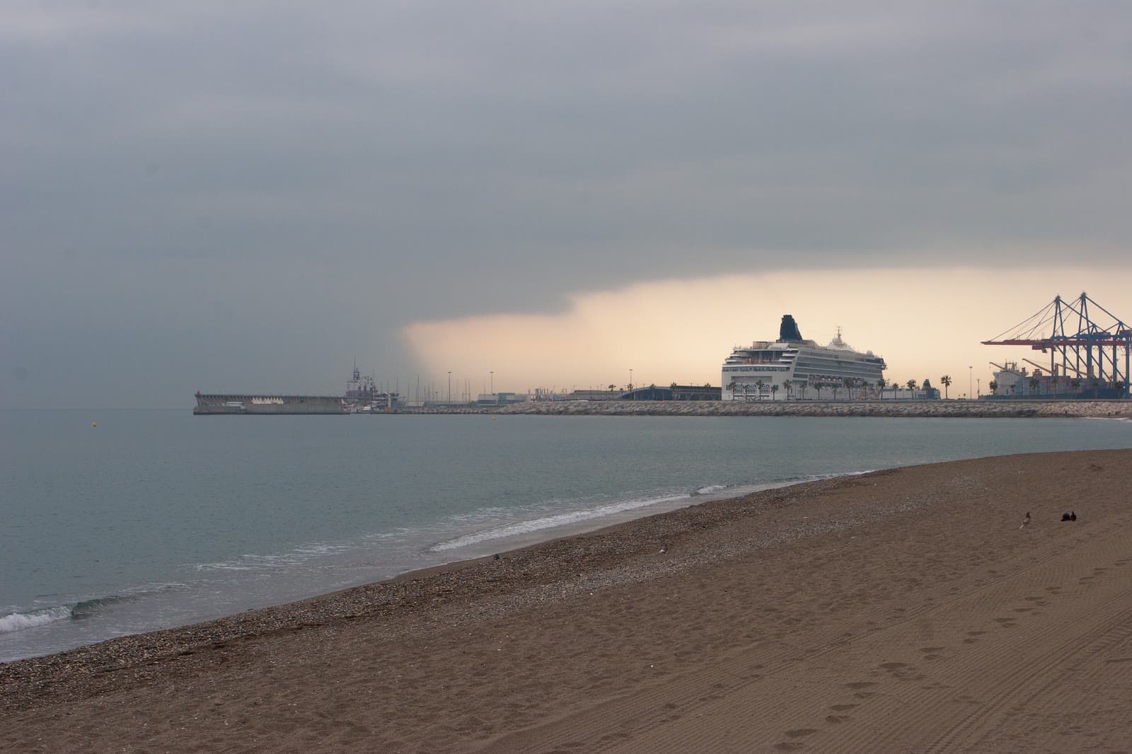 Una cortina de lluvia avanza sobre el puerto de Málaga, con un crucero atracado, vista desde una playa vacía