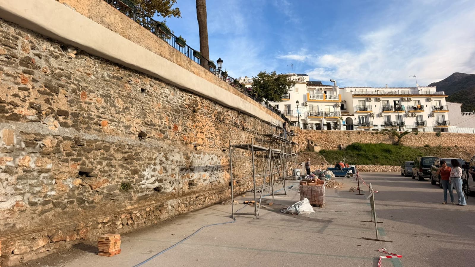 Muro de piedra en la plaza de Maro, con andamios, palés de ladrillo y vallas de obra en el aparcamiento, con edificios blancos al fondo