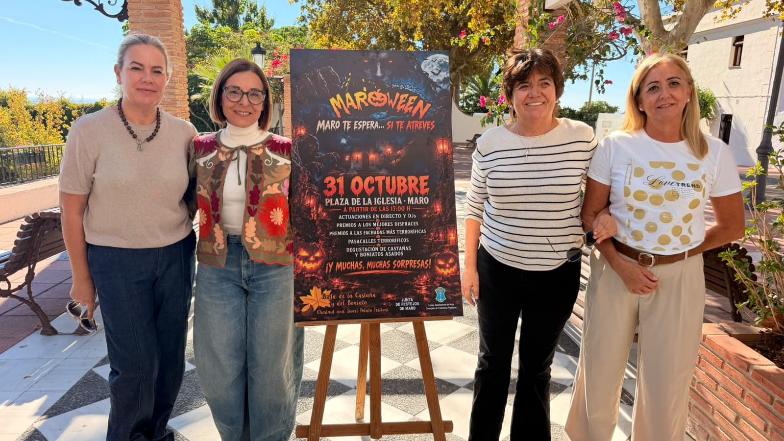 Cuatro mujeres posan en una plaza de Maro junto a un caballete con el cartel de Maroween, que anuncia la fiesta el 31 de octubre en la Plaza de la Iglesia a partir de las 17:00.