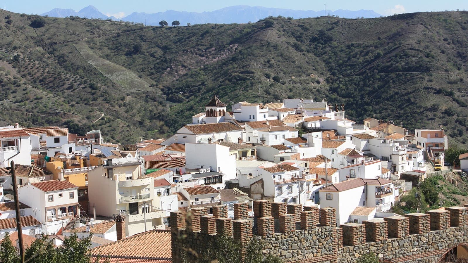 Vista de Moclinejo, con sus casas blancas y tejados rojos y la torre de la iglesia, en una ladera entre montes verdes.