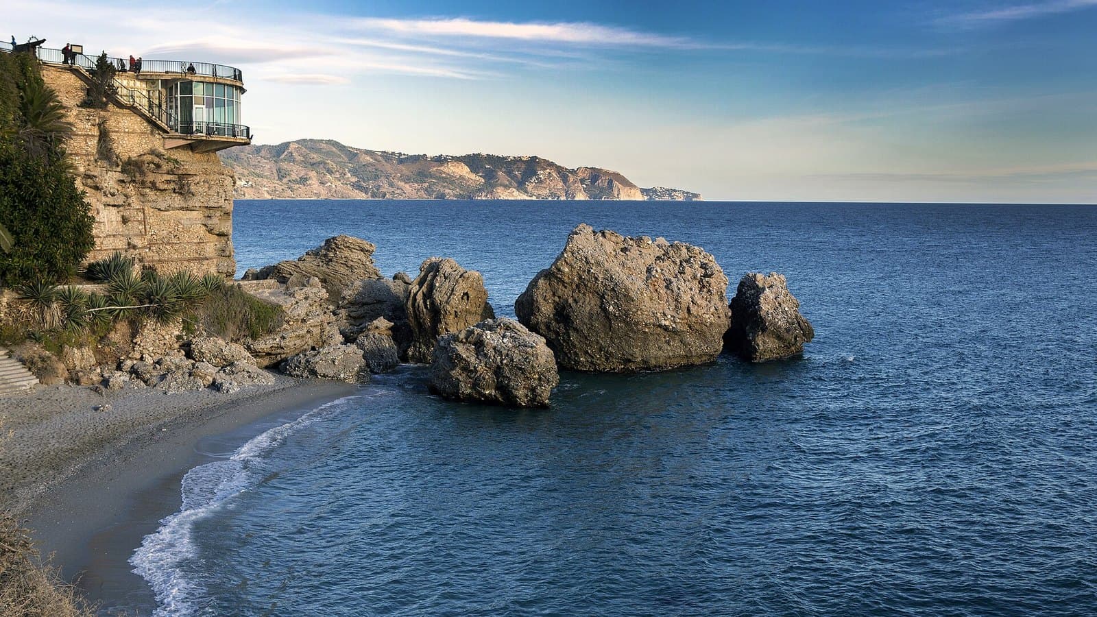 El Balcón de Europa de Nerja sobre la playa de la Calahonda, con grandes rocas en el mar y la costa de la Axarquía al fondo bajo un cielo azul.