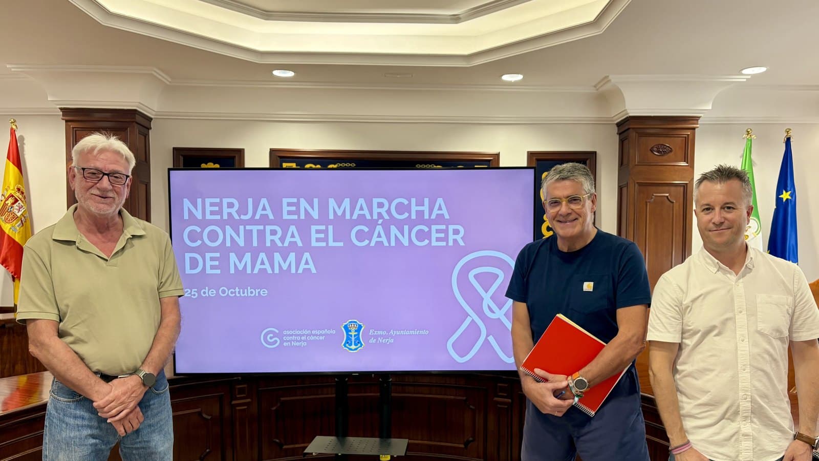 Tres hombres posan en el salón de plenos del Ayuntamiento de Nerja junto a una pantalla que anuncia «Nerja en marcha contra el cáncer de mama, 25 de octubre» con un lazo