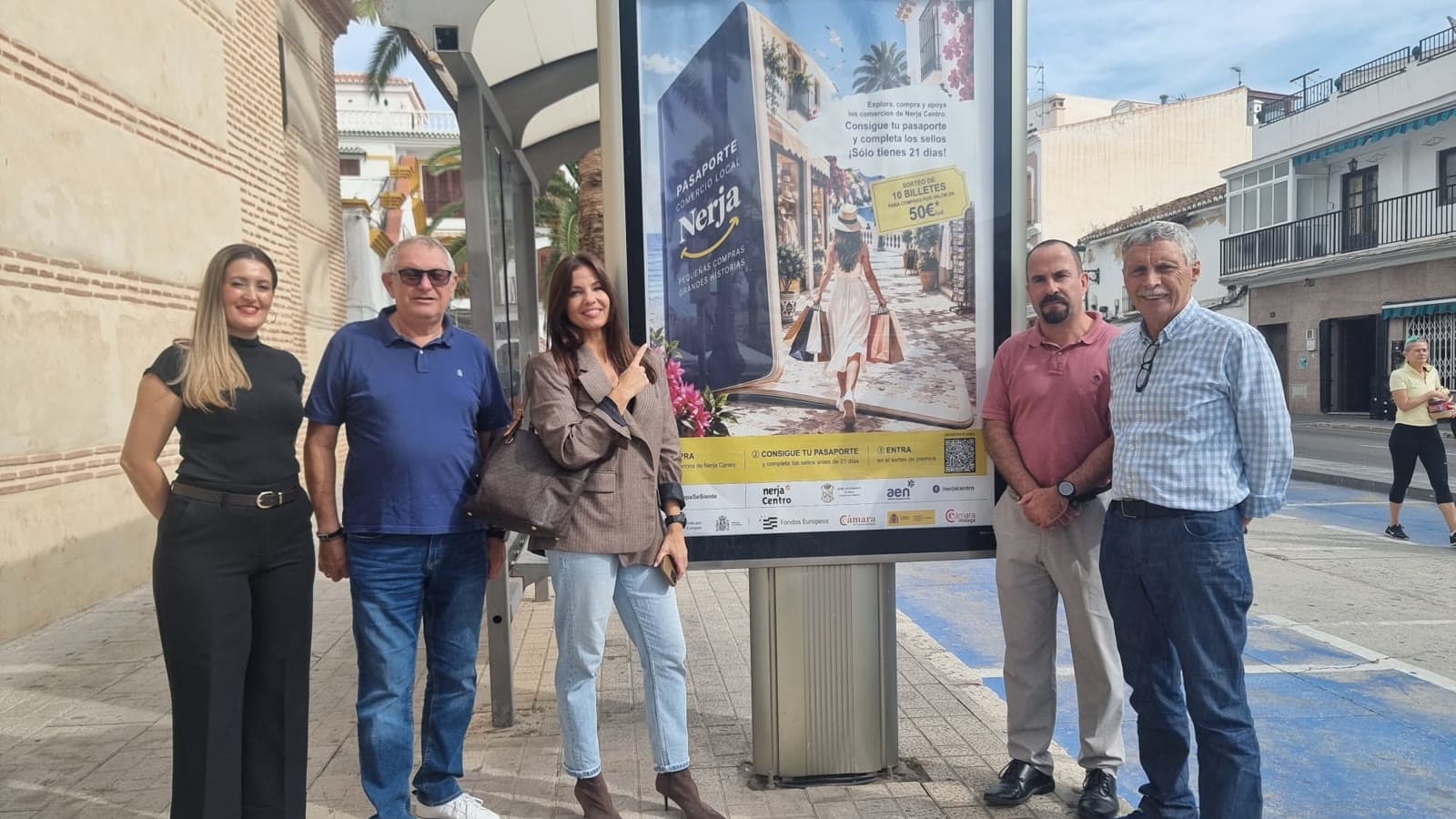 Cinco personas posan en una acera de Nerja junto a una marquesina con el cartel de la campaña «Pasaporte Comercio Local Nerja», que anuncia el sorteo de 10 billetes de 50 euros