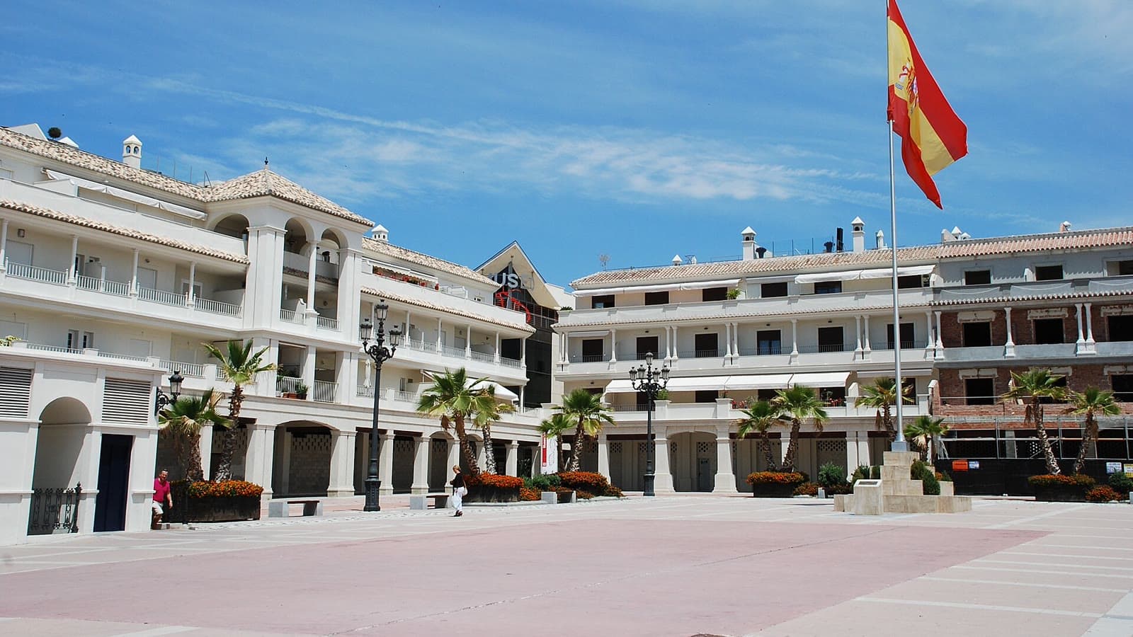 Plaza de España de Nerja, amplia explanada de losas rosadas rodeada de edificios blancos con soportales y palmeras, con una bandera de España en un mástil