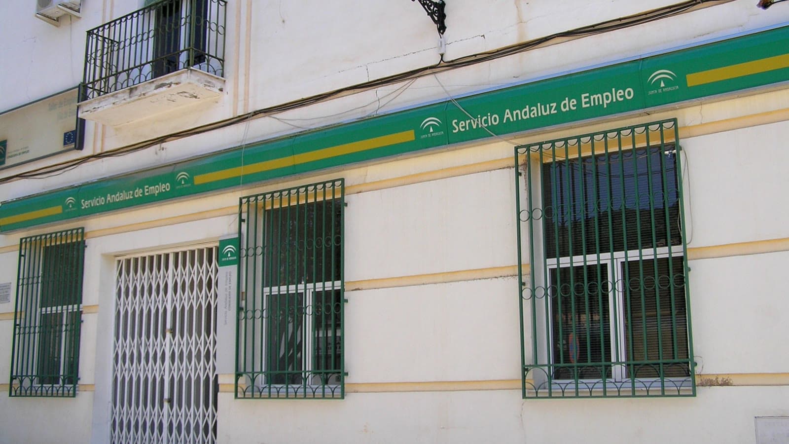 Fachada encalada de una oficina del Servicio Andaluz de Empleo, con un rótulo verde y amarillo de la Junta de Andalucía, ventanas con rejas verdes y una puerta de reja plegable.