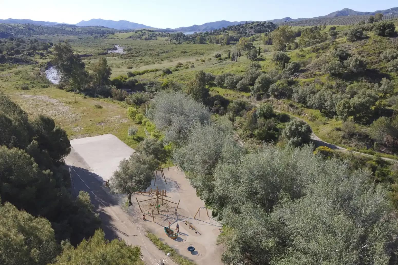 Vista aérea de Las Mayoralas: un parque infantil de juegos de madera sobre una explanada de tierra clara rodeada de olivos, con el valle verde, el cauce del río y, al fondo, el embalse de La Viñuela entre montañas