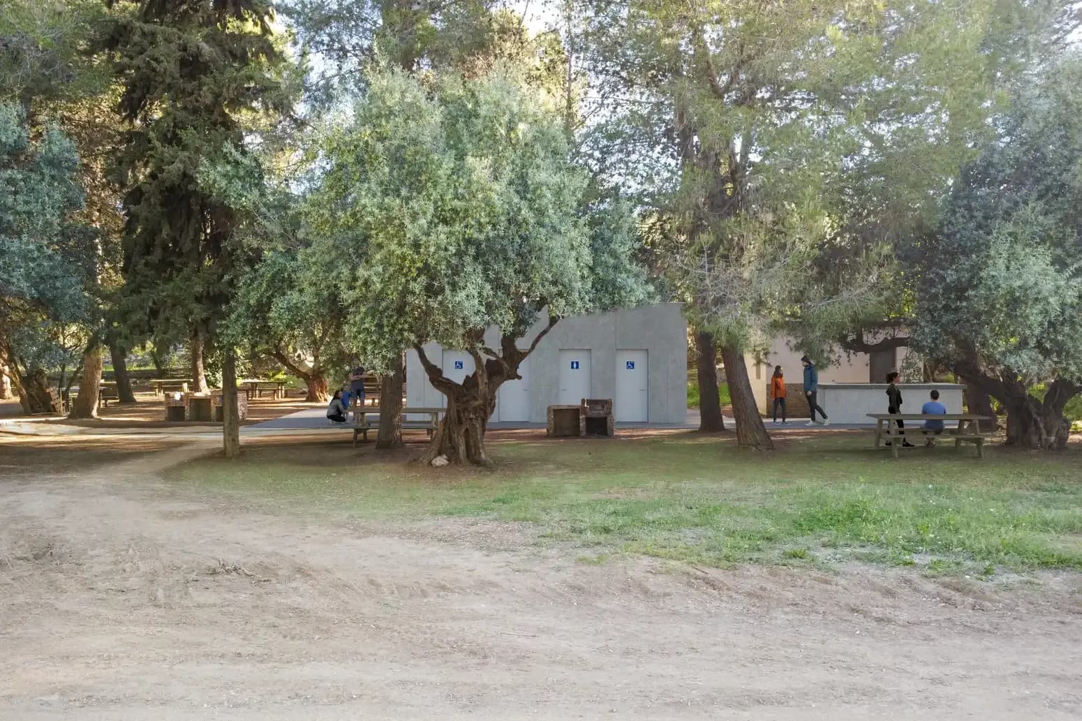 Recreación de la zona de servicios de Las Mayoralas: un edificio gris de una planta con puertas de aseos, accesibles y de caballeros, entre olivos y pinos, con mesas de pícnic y barbacoas de obra delante