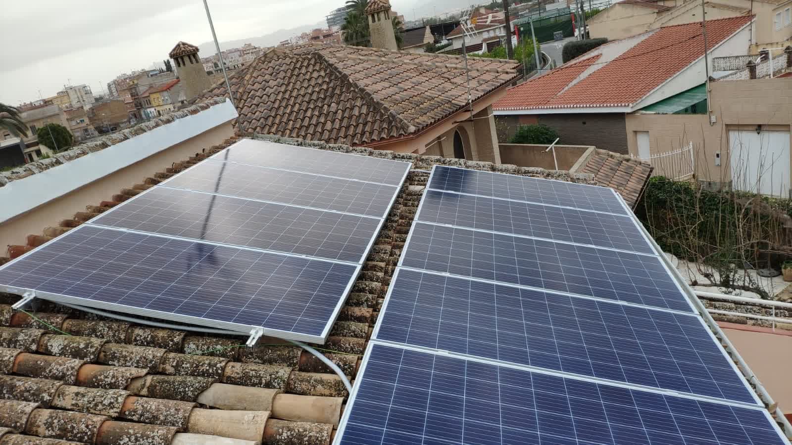 Placas solares fotovoltaicas instaladas sobre el tejado de teja de una vivienda, con otras casas al fondo.