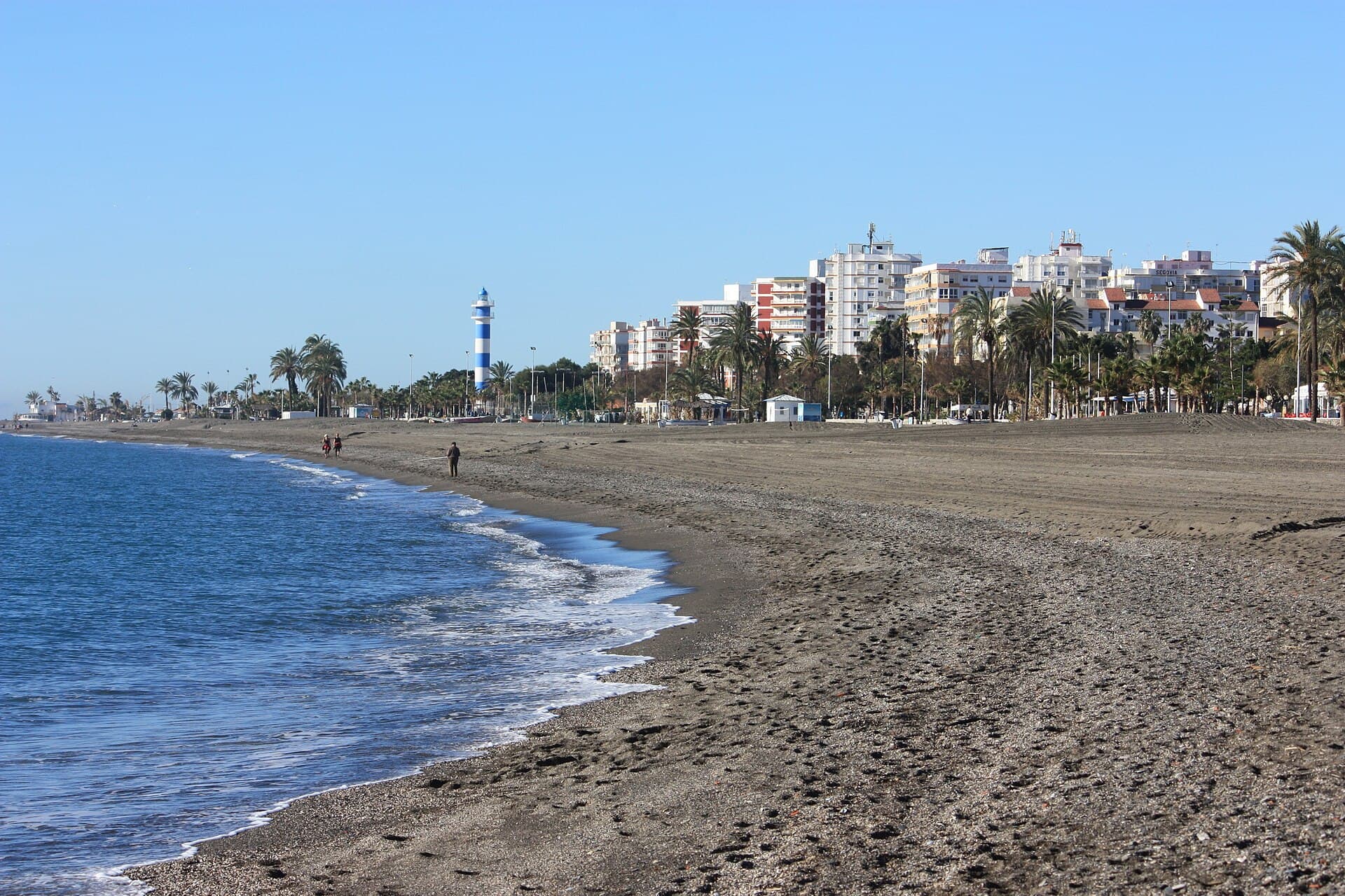 Playa de Torre del Mar con el faro y los edificios del paseo marítimo al fondo, bajo un cielo despejado.