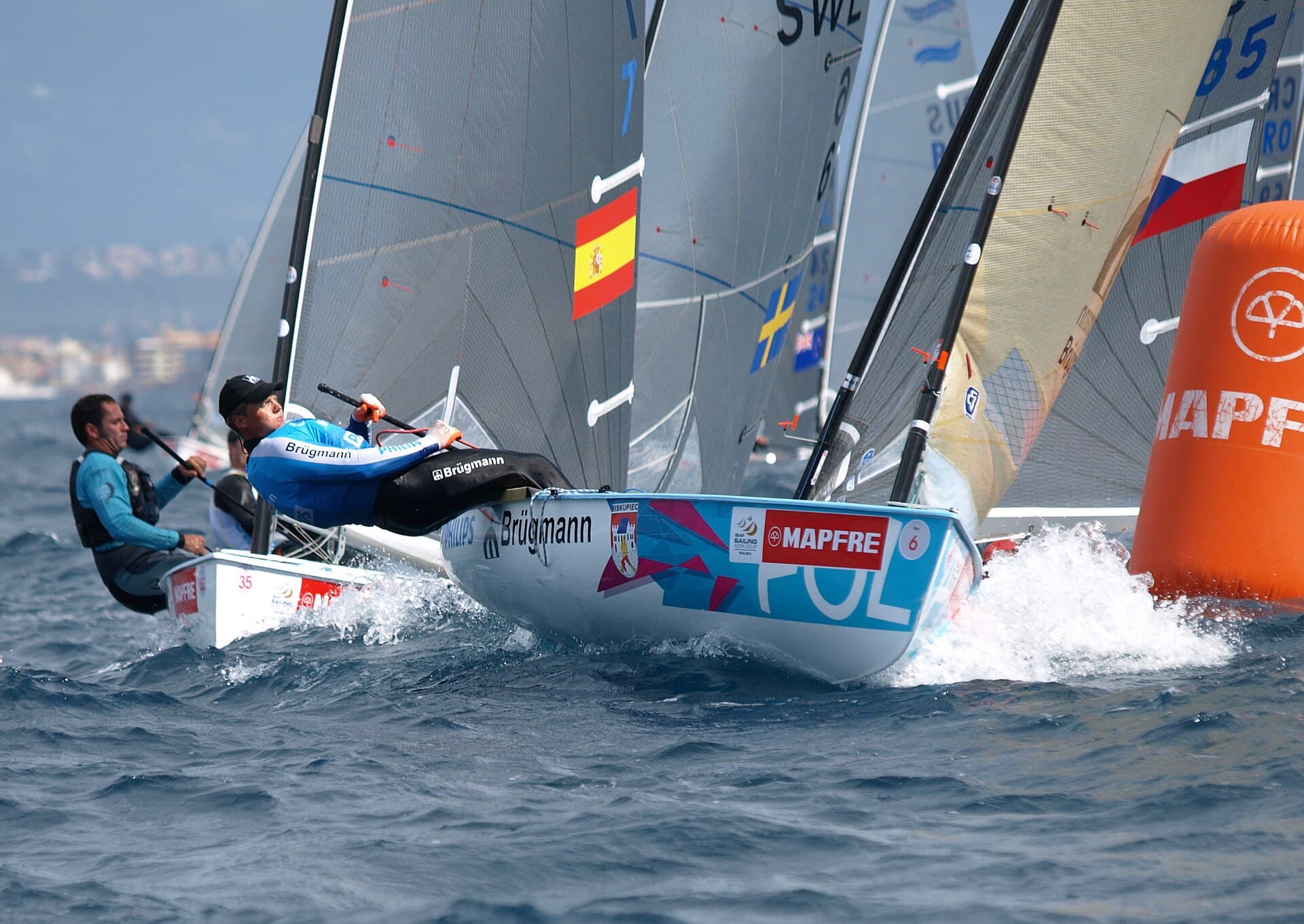 Dos veleros de la clase Finn navegan escorados en una regata, con el agua salpicando y una boya naranja al fondo.