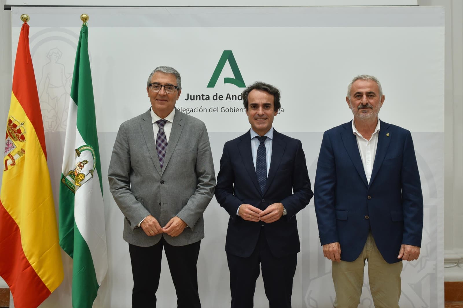 Tres hombres con chaqueta posan de pie ante un panel blanco con el logotipo de la Junta de Andalucía, junto a las banderas de España y Andalucía.