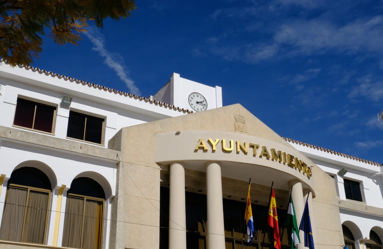 Fachada del Ayuntamiento de Rincón de la Victoria, con el rótulo «Ayuntamiento» en letras doradas, columnas, banderas y la torre del reloj bajo un cielo azul