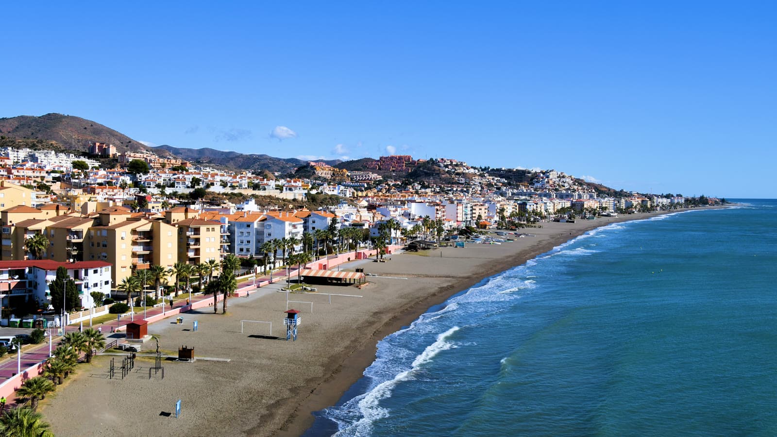 Vista desde lo alto de la playa y el paseo marítimo de Rincón de la Victoria, con bloques de pisos junto a la arena y lomas con urbanizaciones al fondo.