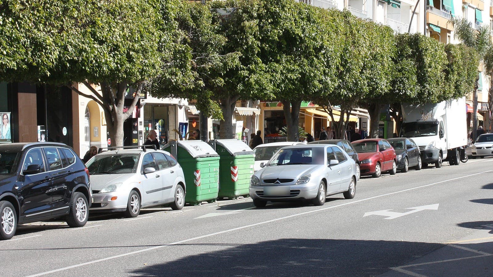 Coches aparcados en línea bajo los árboles del Paseo de la Marina, en Rincón de la Victoria, con comercios y edificios de viviendas al fondo.