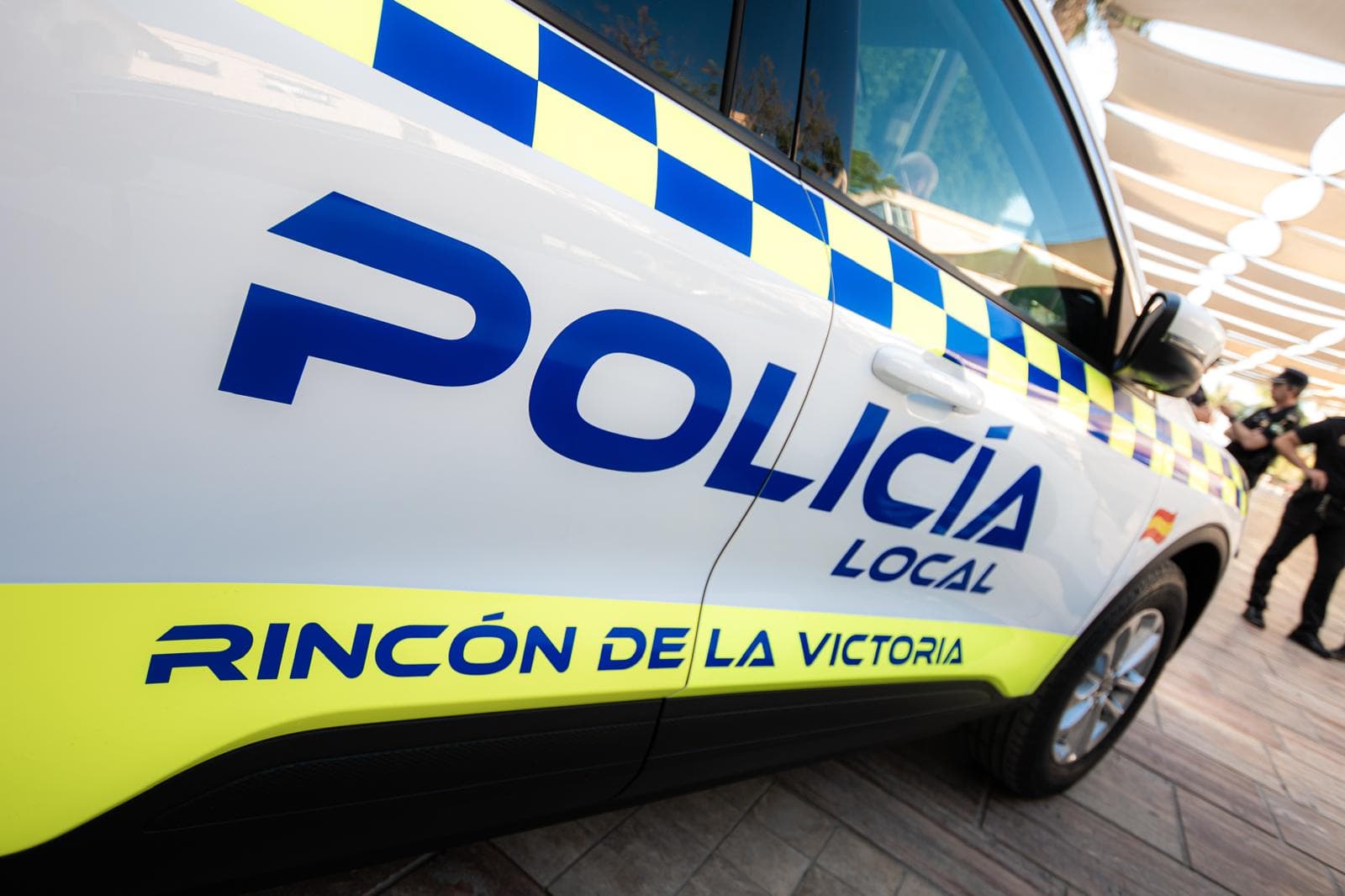 Lateral de un coche patrulla de la Policía Local de Rincón de la Victoria, con el rótulo «Policía Local» y la banda azul y amarilla, y dos agentes al fondo.