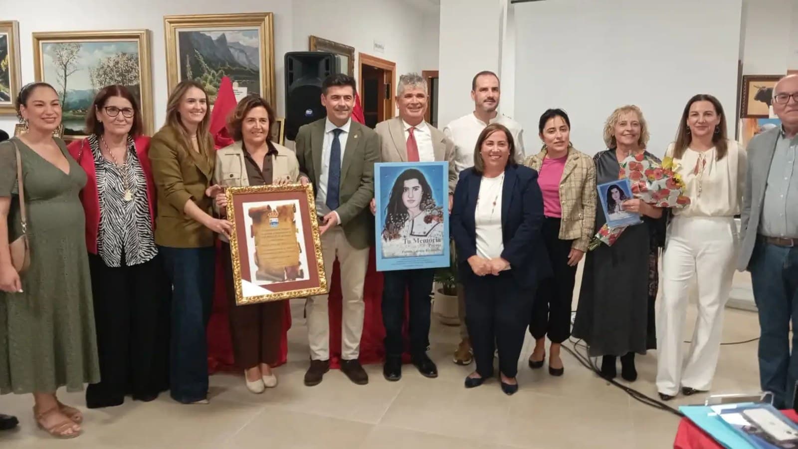 Una docena de personas posan en una sala con cuadros de paisajes en las paredes; en el centro sostienen un diploma enmarcado en dorado y un cartel azul con el retrato de una joven de pelo oscuro y el título «Tu memoria».