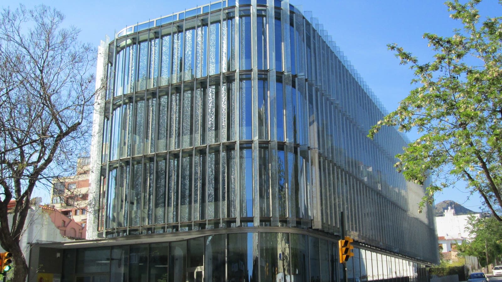 Edificio de oficinas de la Seguridad Social en Málaga, con fachada curva de cristal y lamas metálicas verticales, junto a una calle con árboles y un semáforo.