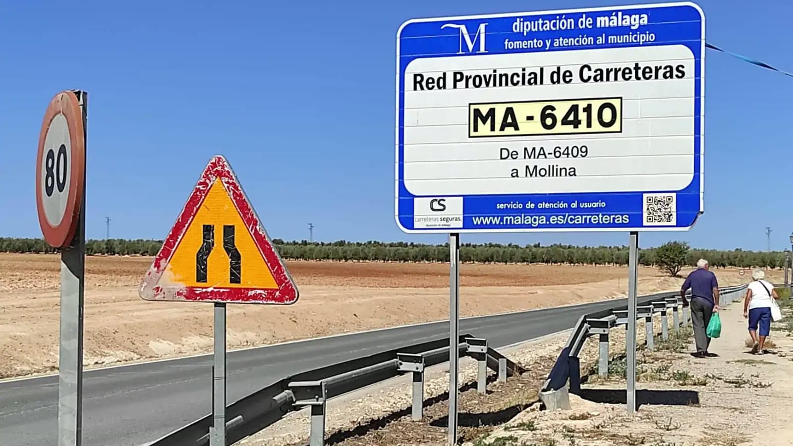 Cartel azul y blanco de la Red Provincial de Carreteras de la Diputación de Málaga junto a una señal de 80 y otra de estrechamiento de calzada, con la pintura desgastada, al borde de una carretera entre campos de olivos.