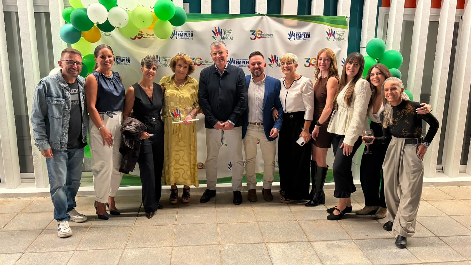 Un grupo de una docena de personas posa ante un photocall con los logotipos del 30 aniversario de la Asociación Taller de la Amistad y de su Centro Especial de Empleo, decorado con globos verdes y blancos.