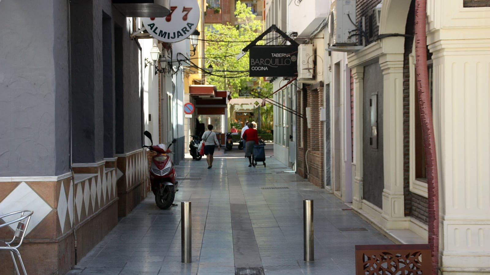 Calle peatonal del centro de Torre del Mar con suelo de baldosas, bolardos metálicos, una moto aparcada, rótulos de comercios y dos personas caminando de espaldas al fondo.