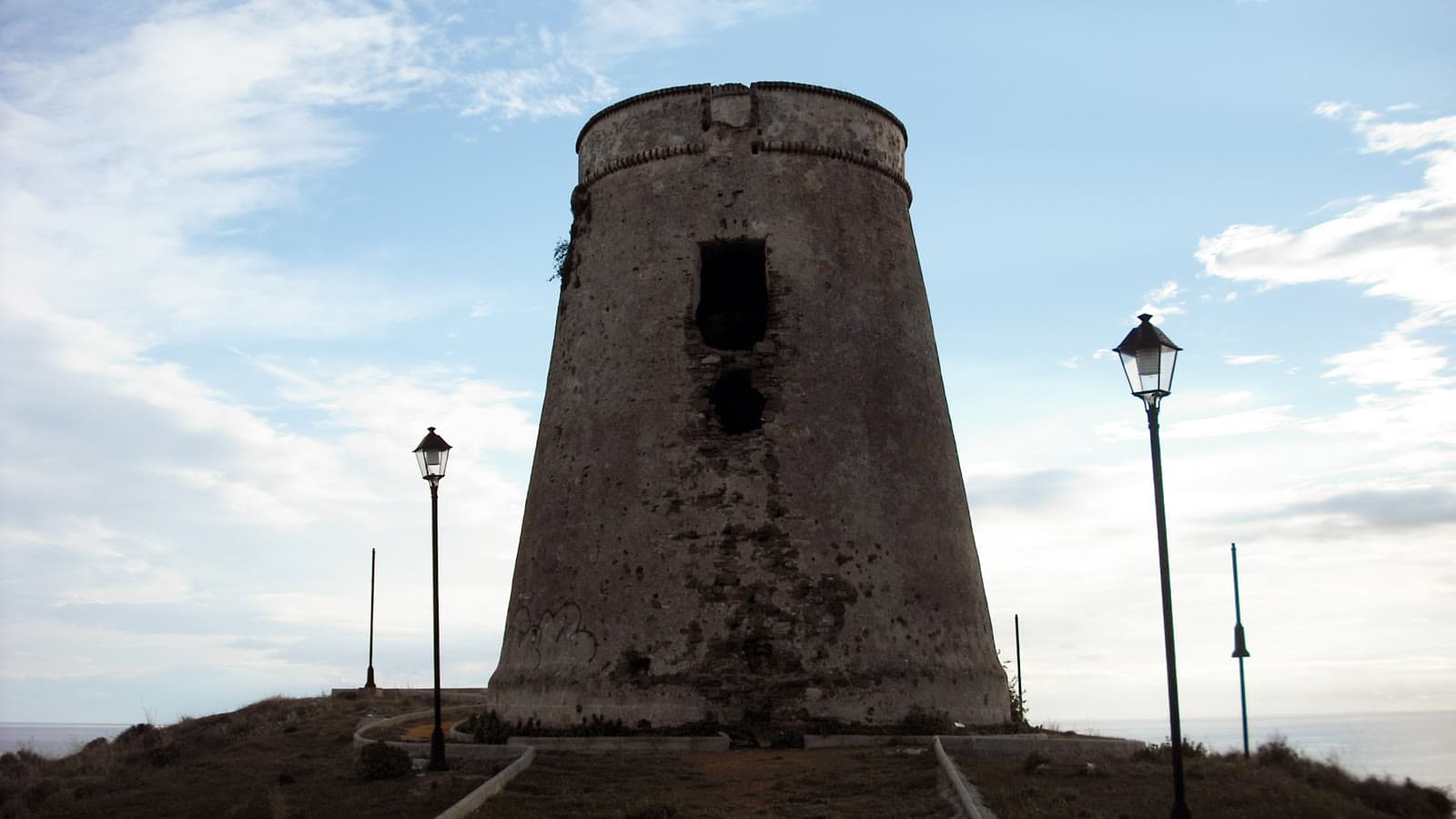Torre de vigilancia troncocónica de piedra, con dos huecos superpuestos en su cara norte, sobre un cerro con farolas y caminos de bordillo, con el mar al fondo bajo un cielo con nubes.